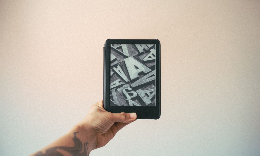 A tattooed hand holding an e-reader displaying typography design against a plain background.