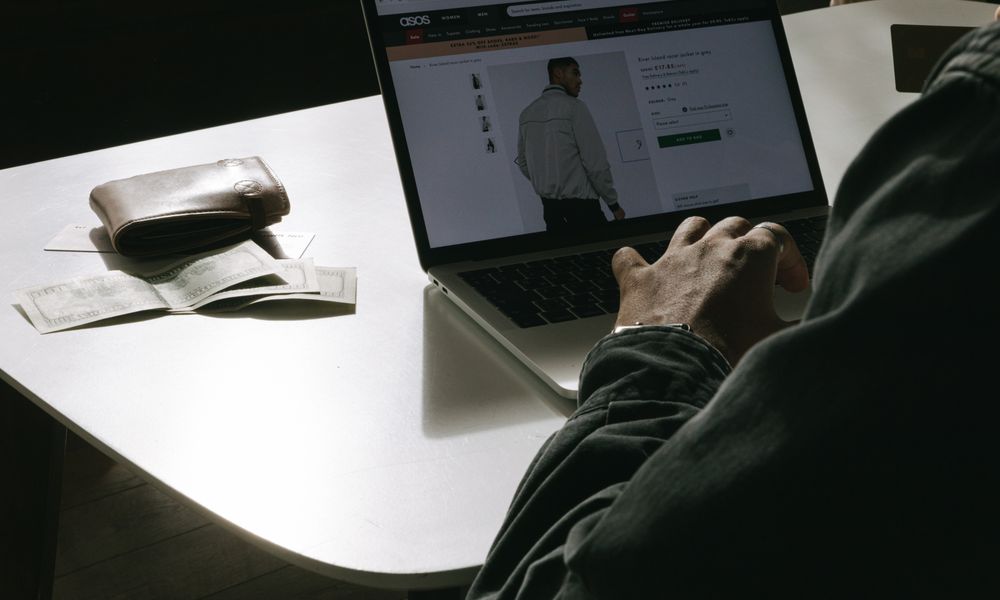 Adult man shopping for clothes online at home using a laptop, with a wallet on the table.