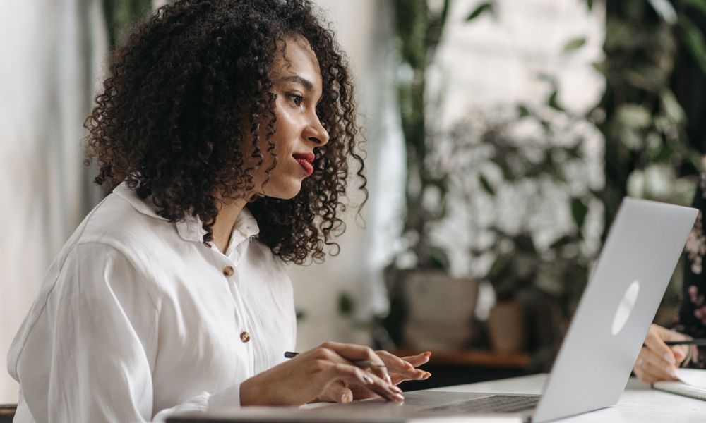 Young woman with curly hair typing on laptop in a bright, plant-filled office space.
