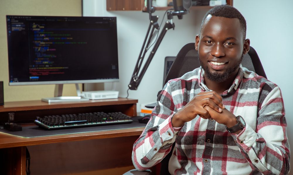 Portrait of a smiling programmer at a desk with computer screen displaying code.