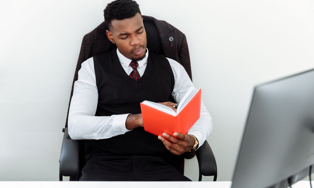 Focused African American man reading a red book at his workspace in an office setting.