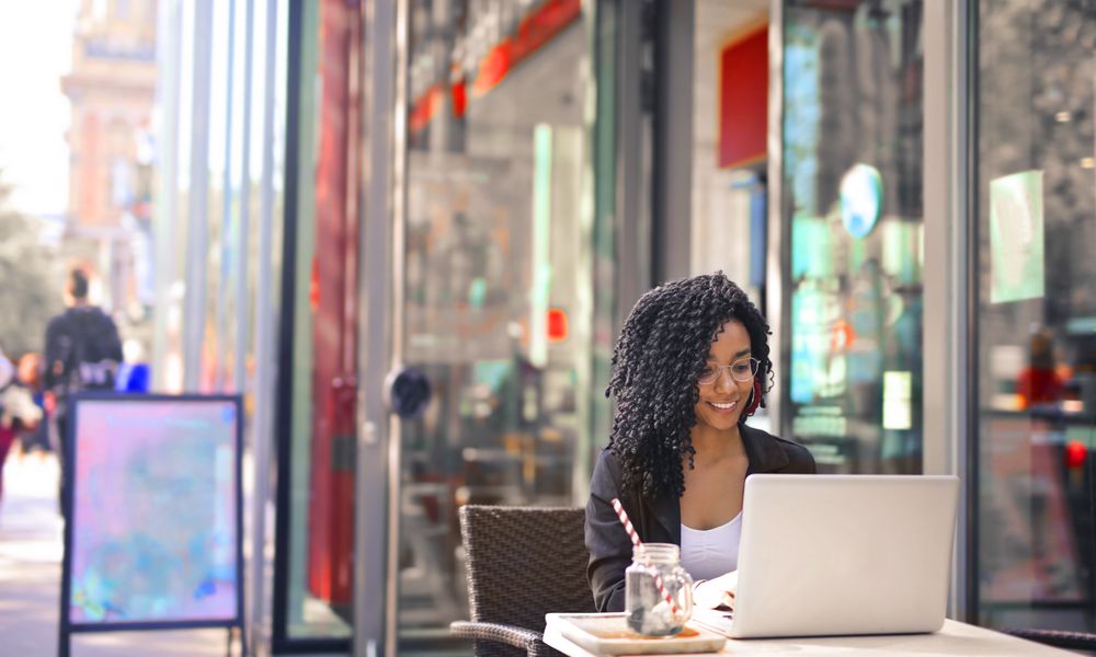 Smiling woman with curly afro hair typing on a laptop outside a modern cafe.