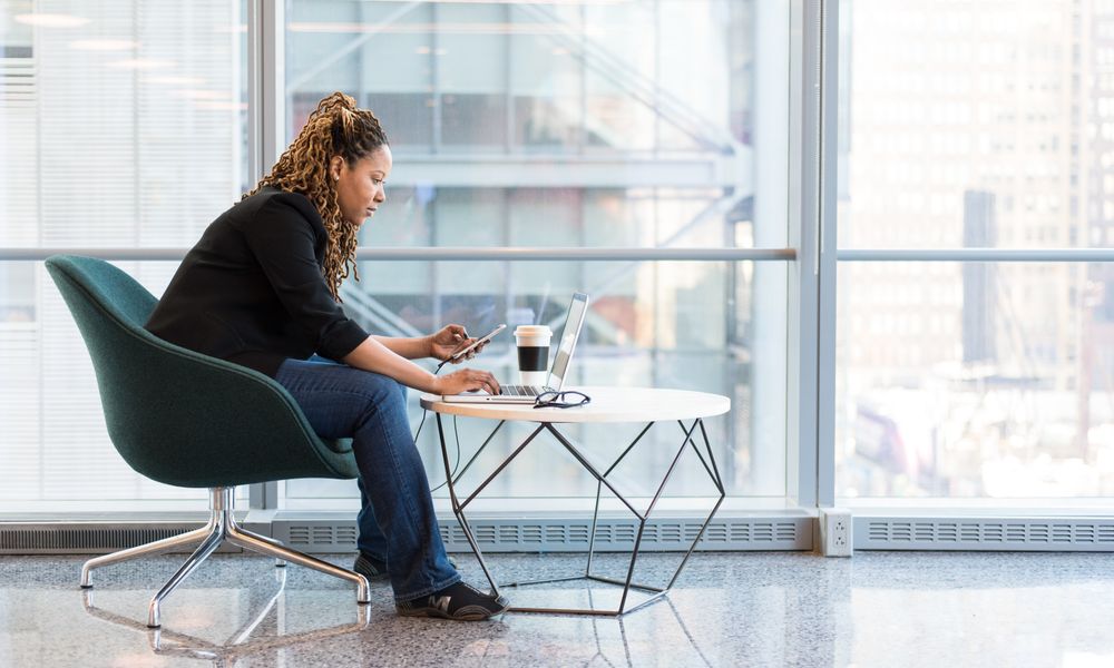 Business woman using smartphone and laptop in a bright, modern office space. Ideal for technology and work themes.