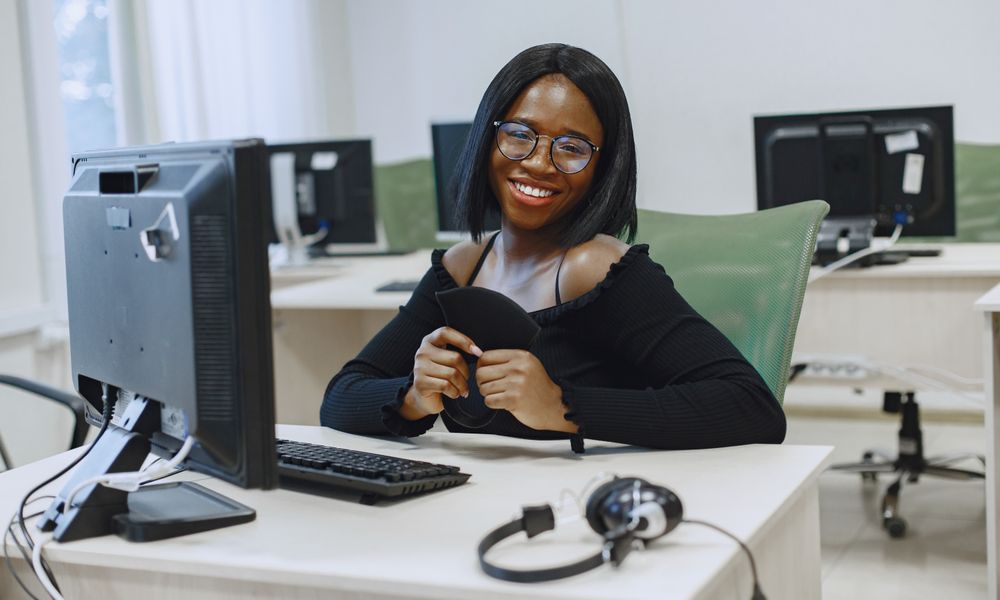 African American woman smiling at a computer desk in an office setting, holding a black item.