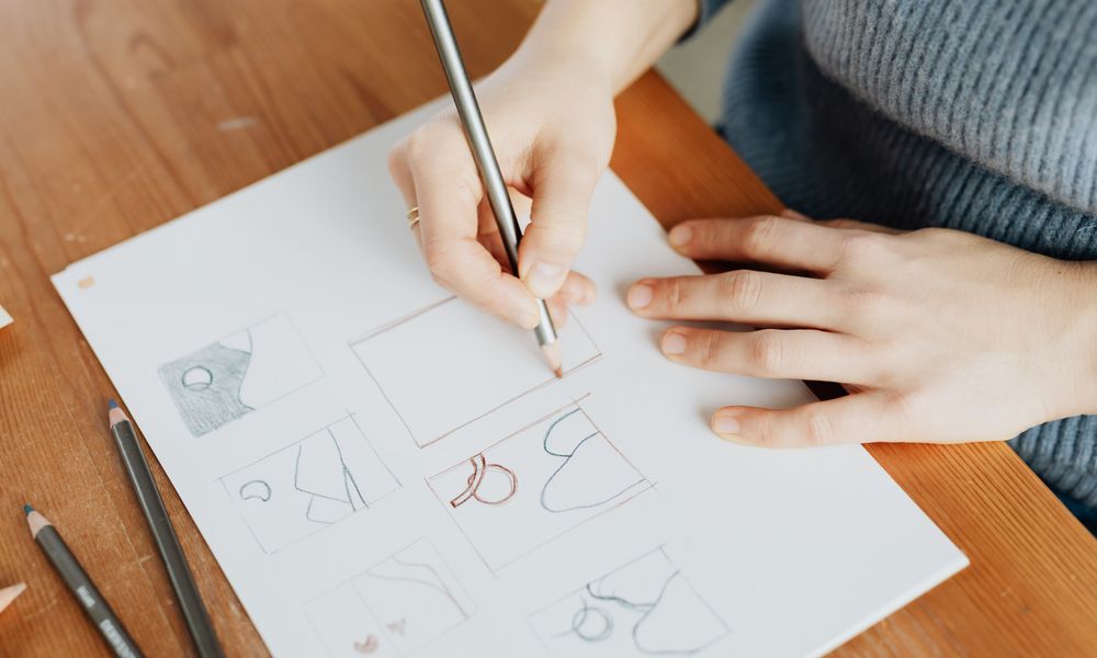 From above of faceless female designer in knitted sweater sitting at wooden table with sheet of paper while drawing with penc