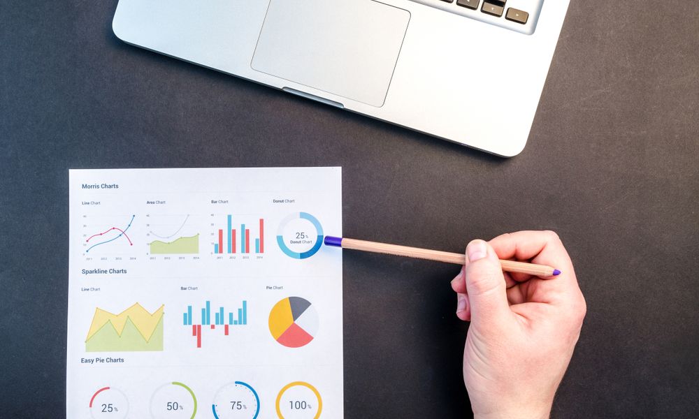 Hand holding pencil reviewing colorful data charts on desk with laptop.