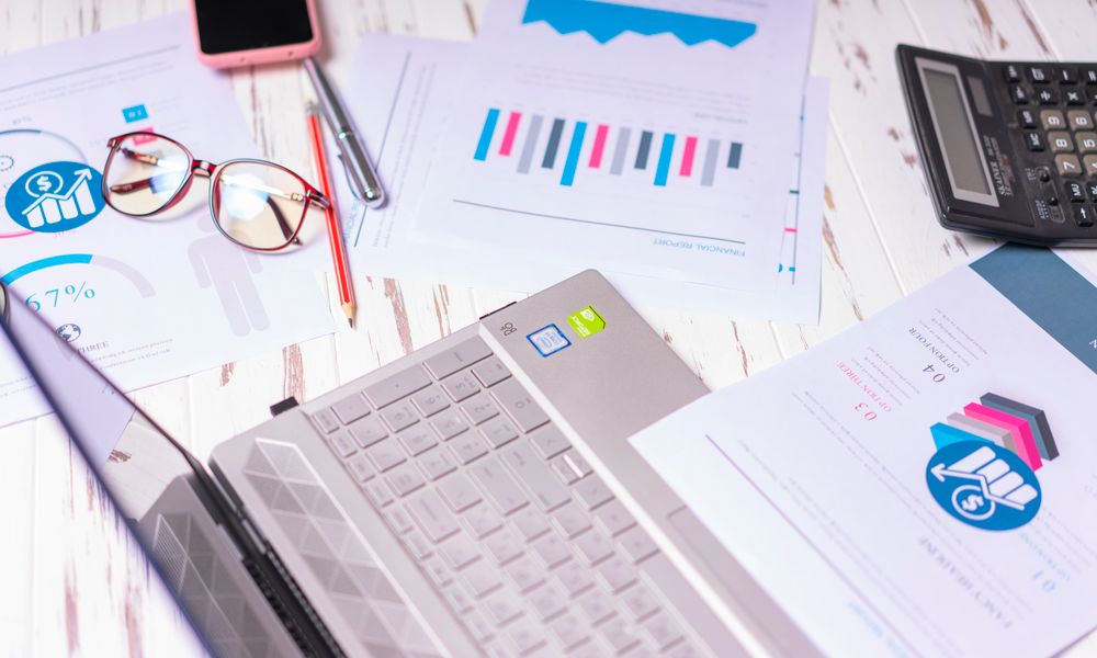A cluttered desk with a laptop, financial reports, calculator, and glasses indicating a busy work environment.