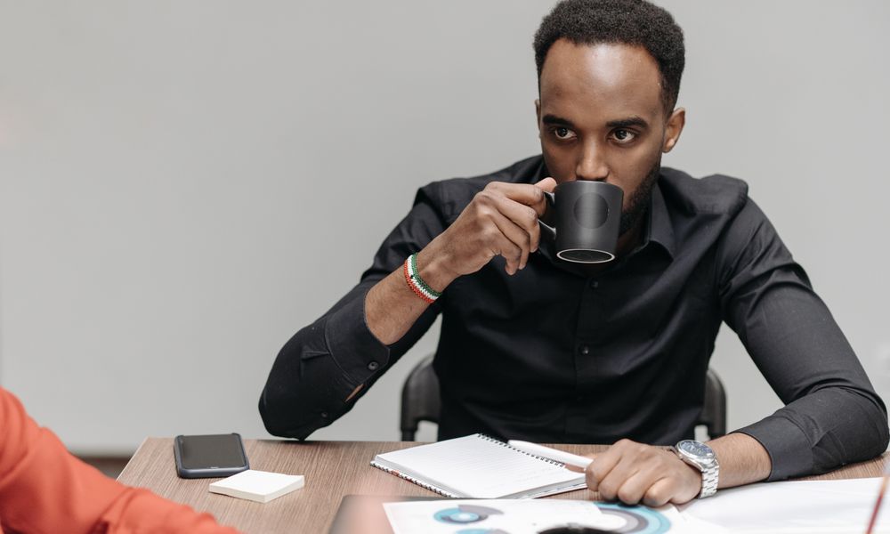 Man in a black shirt drinking coffee while sitting at an office table with documents nearby.