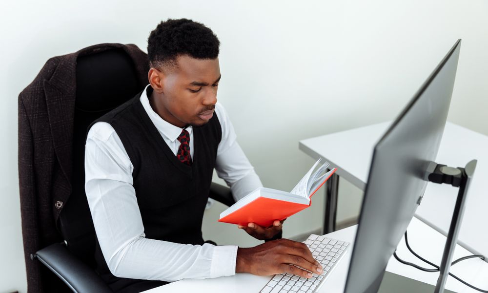 Business professional reading a book while working at the computer in the office.