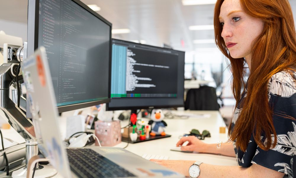 A focused female software engineer coding on dual monitors in a modern office.