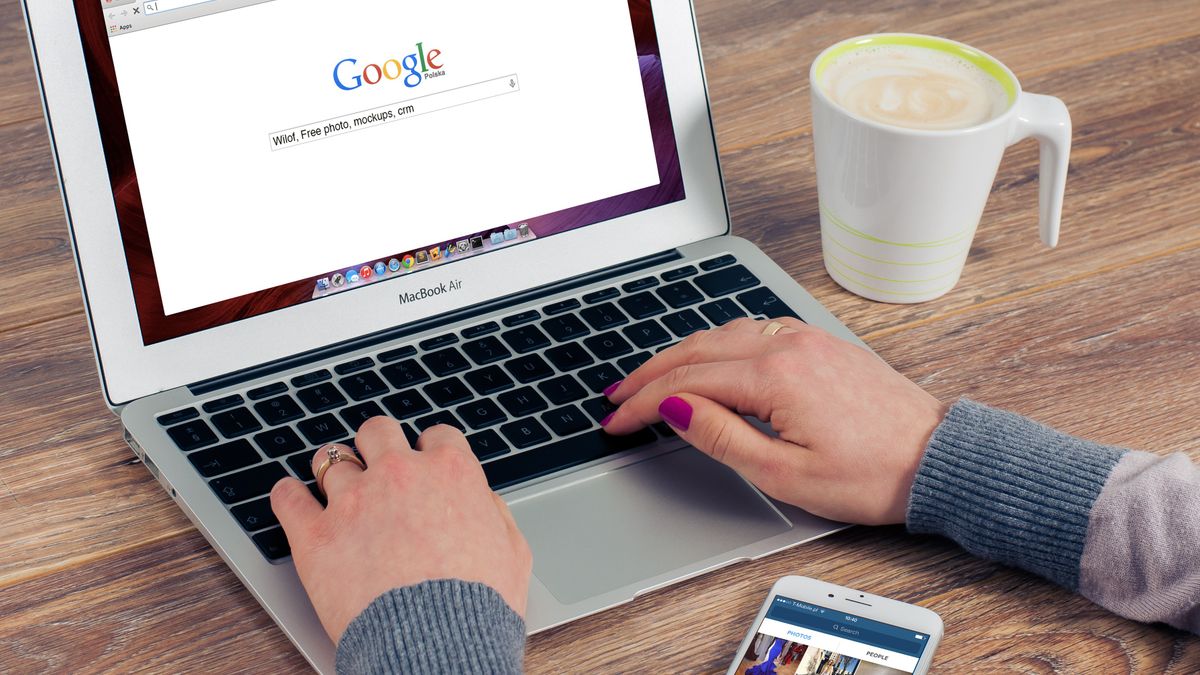 Hands typing on MacBook Air with Google search open, coffee nearby.