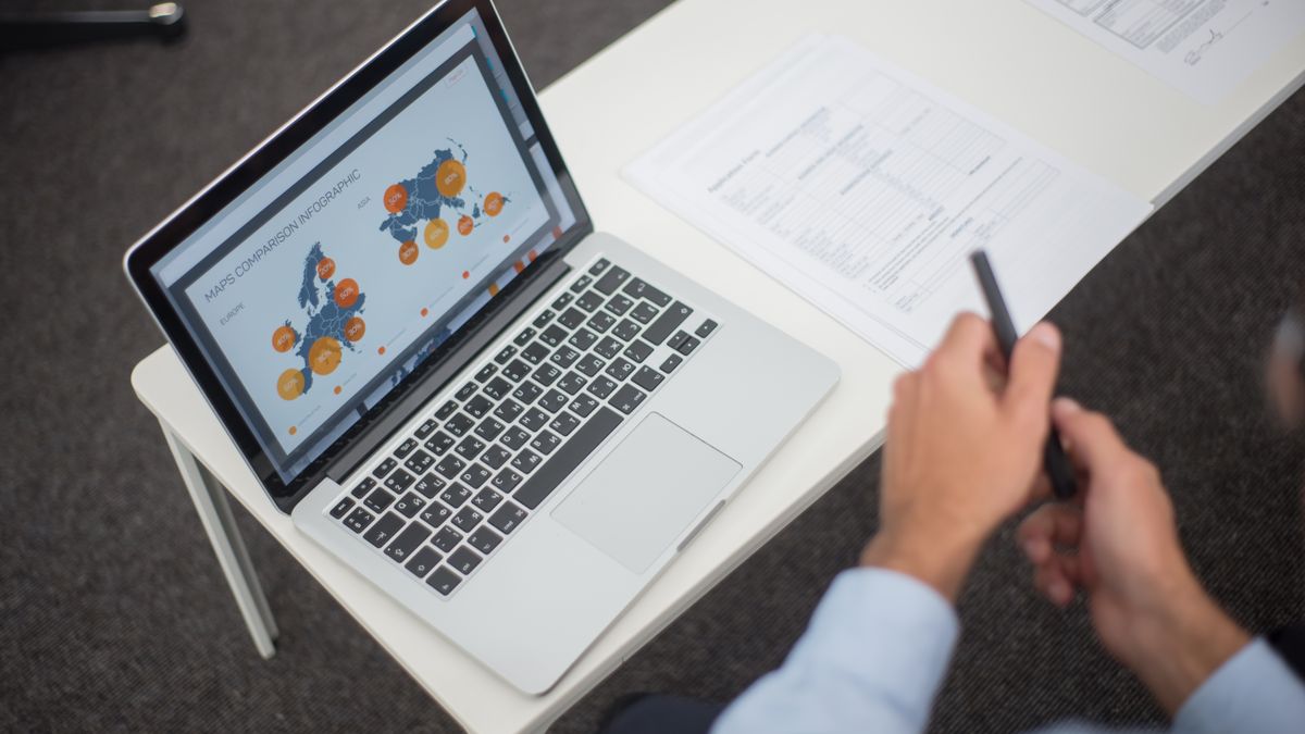 Professional analyzing data infographics on a laptop at an office desk, featuring documents and a pen.