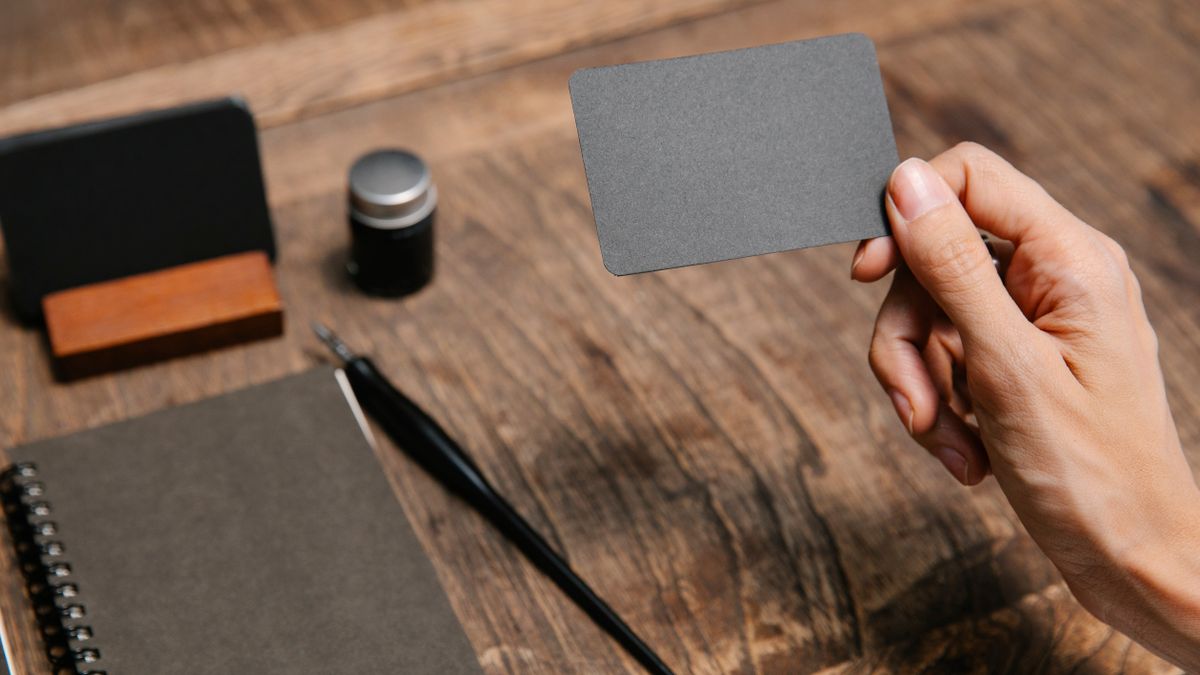 A close-up of a hand holding a blank business card above a wooden desk with office accessories.