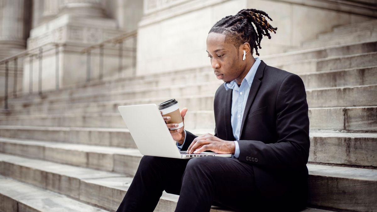 Concentrated African American businessman wearing formal clothes with coffee to go and earbuds browsing netbook while working remotely