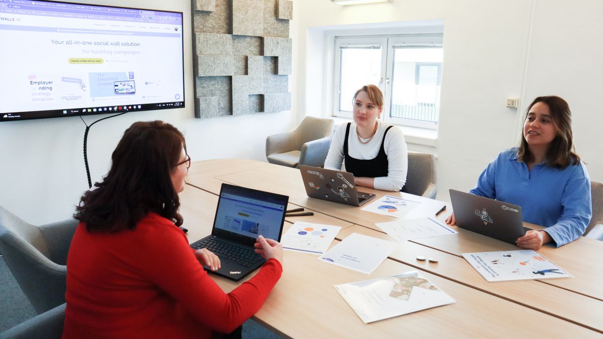 Three women collaborating at a conference table in a modern office with laptops and presentation screen.